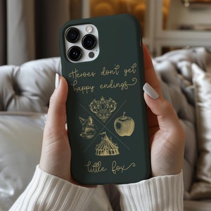 May include: A dark green phone case with a gold design. The case features a quote that reads "Heroes don't get happy endings." and a graphic of a fox, apple, heart, and tent. The text "little fox" is at the bottom of the case.