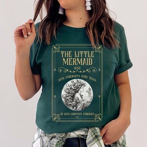 Pu&ograve; includere: Una maglietta verde con una grafica dorata di una copertina di libro con il testo "The Little Mermaid", "Hans Andersen's Fairy Tales", e "By Hans Christian Andersen 1843".