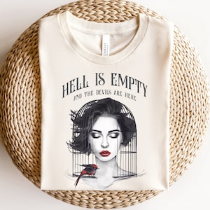 Puede incluir: Una camiseta beige con el texto "El infierno está vacío y los demonios están aquí" impreso en negro. Una ilustración en blanco y negro de una mujer con los ojos cerrados y un pájaro rojo en una jaula está impresa en la camiseta.