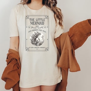 Pu&ograve; includere: Una maglietta bianca con un'illustrazione vintage di una sirena con il testo "The Little Mermaid" e "Hans Andersen's Fairy Tales" in una cornice nera.