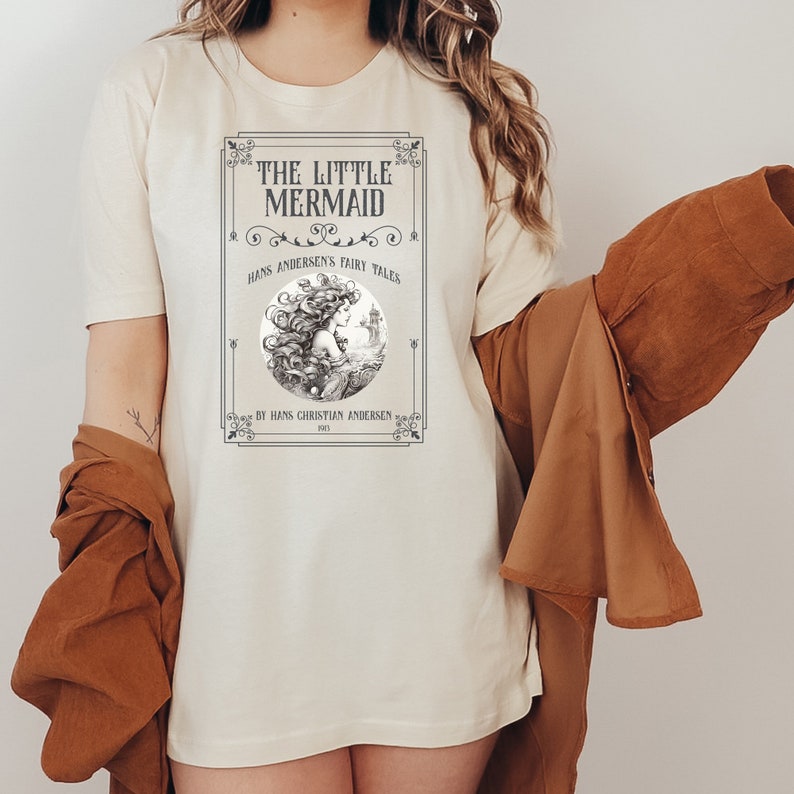 Pu&ograve; includere: Una maglietta bianca con un'illustrazione vintage di una sirena con il testo "The Little Mermaid" e "Hans Andersen's Fairy Tales" in una cornice nera.