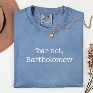 May include: A light blue Comfort Colors t-shirt with the text "fear not, Bartholomew" in white. A gold necklace with a star pendant is draped across the shirt. A brown hat and flowers are in the background.