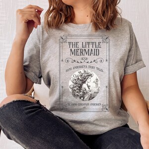 Pu&ograve; includere: Maglietta grigia con un design di copertina di libro vintage con un'illustrazione di sirena e il testo "The Little Mermaid, Hans Andersen's Fairy Tales, By Hans Christian Andersen 1837".