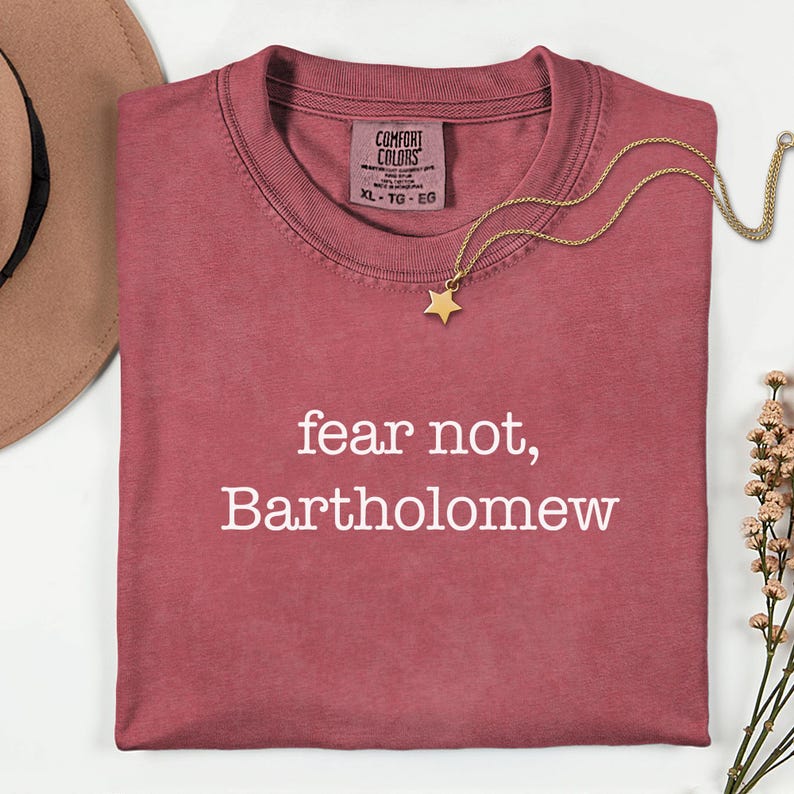 May include: A maroon Comfort Colors t-shirt with the text "fear not, Bartholomew" in white. A gold necklace with a star pendant rests on the shirt. A brown hat and sprig of flowers are in the background.
