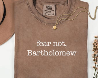 May include: A brown Comfort Colors t-shirt with the text "fear not, Bartholomew" in white. A gold star pendant necklace and a brown hat are also visible. The shirt is size XL-TG-EG.
