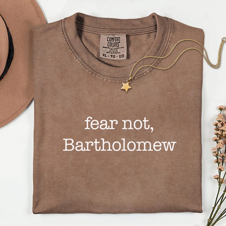 May include: A brown Comfort Colors t-shirt with the text "fear not, Bartholomew" in white. A gold star pendant necklace and a brown hat are also visible. The shirt is size XL-TG-EG.