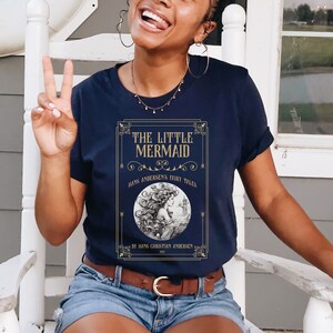 Pu&ograve; includere: Una t-shirt blu navy con una grafica dorata di una copertina di libro con il testo "The Little Mermaid" e un'illustrazione di una sirena. Il testo "Hans Andersen's Fairy Tales" e "By Hans Christian Andersen 1837" sono inclusi anche sulla copertina.