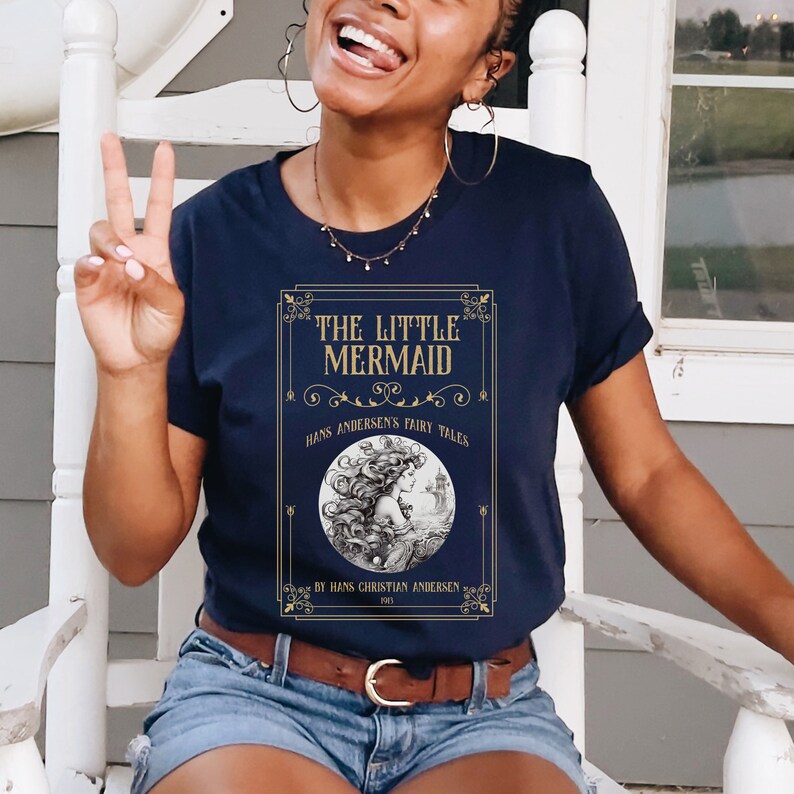 Pu&ograve; includere: Una t-shirt blu navy con una grafica dorata di una copertina di libro con il testo "The Little Mermaid" e un'illustrazione di una sirena. Il testo "Hans Andersen's Fairy Tales" e "By Hans Christian Andersen 1837" sono inclusi anche sulla copertina.