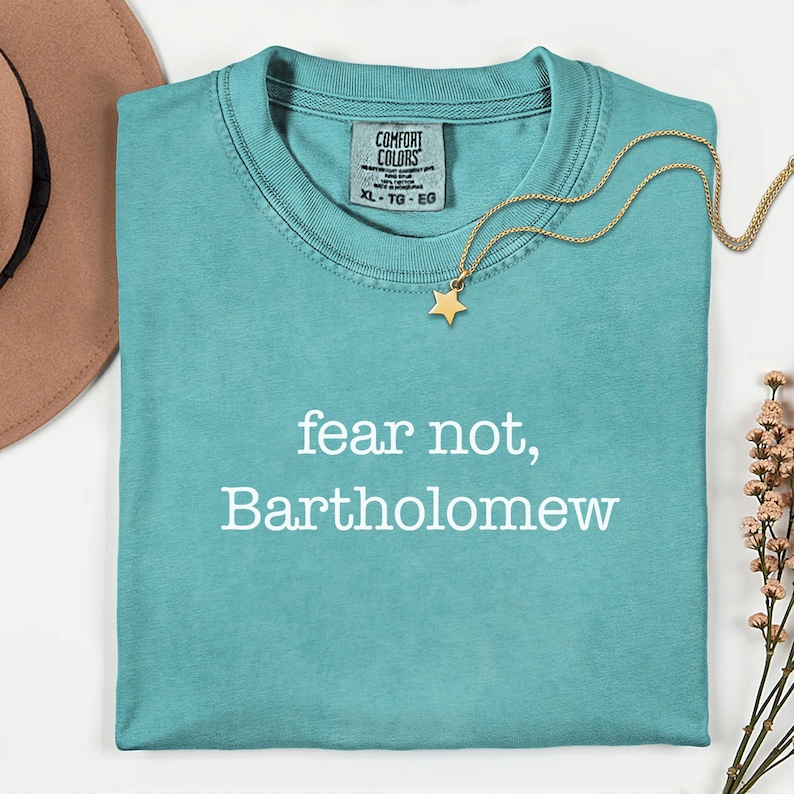 May include: A turquoise Comfort Colors t-shirt with the text "fear not, Bartholomew" in white. A gold star pendant necklace and a brown hat are also in the image. The shirt is XL-TG-EG.