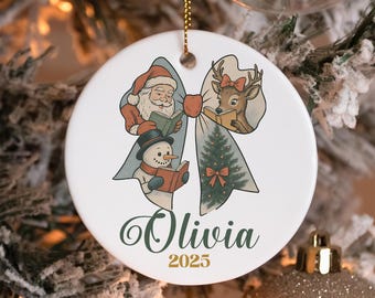 May include: A white ceramic Christmas ornament with a festive design. It features Santa Claus, a reindeer, and a snowman reading books, set against a bow-shaped backdrop. The name "Olivia" and the year "2025" are also displayed.