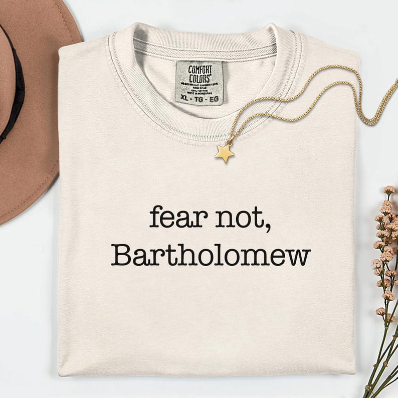 May include: Cream-colored t-shirt with the text "fear not, Bartholomew" in black. A gold necklace with a star pendant and a brown hat are also visible. The shirt has a Comfort Colors tag.