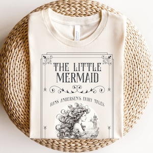 Pu&ograve; includere: Una t-shirt beige con una stampa grafica in bianco e nero di una sirena con il testo "The Little Mermaid" e "Hans Andersen's Fairy Tales".