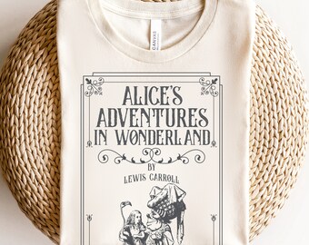 May include: A cream-colored t-shirt with a vintage-style book cover design featuring the text "Alice's Adventures in Wonderland" by Lewis Carroll. The cover includes an illustration of Alice and the Mad Hatter.