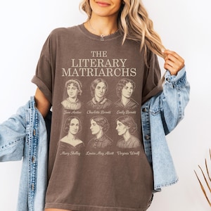 Puede incluir: Camiseta marrón con el texto "THE LITERARY MATRIARCHS" y retratos de Jane Austen, Charlotte Brontë, Emily Brontë, Mary Shelley, Louisa May Alcott y Virginia Woolf. La camiseta tiene mangas cortas.