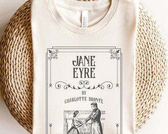 Pode incluir: Uma camiseta de cor creme com um gráfico preto e branco da capa do livro "Jane Eyre" de Charlotte Brontë. O gráfico mostra um casal em pé em frente a uma lareira.