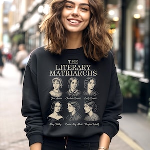 Puede incluir: Sudadera negra con el texto "THE LITERARY MATRIARCHS" y retratos de Jane Austen, Charlotte Brontë, Emily Brontë, Mary Shelley, Louisa May Alcott y Virginia Woolf. Cuello redondo.