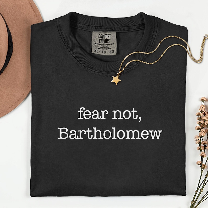 May include: Black Comfort Colors t-shirt with the text "fear not, Bartholomew" in white. A gold star pendant necklace and a brown hat are also visible. The shirt is size XL-TG-EG.