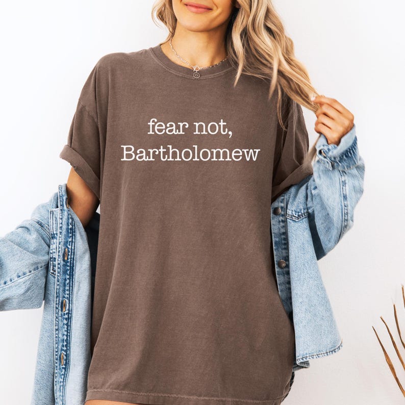 May include: A brown t-shirt with the text "fear not, Bartholomew" in white. The shirt has short sleeves and a relaxed fit. A denim jacket is draped over the shoulders.