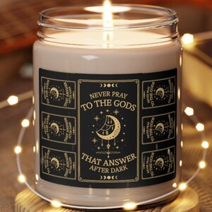 May include: A black and gold candle with a label that reads "Never pray to the gods that answer after dark". The label features a crescent moon with stars and a repeating pattern of the same text.