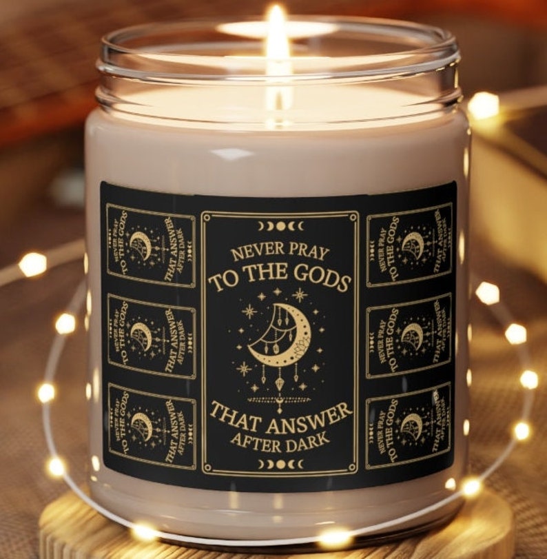 May include: A black and gold candle with a label that reads "Never pray to the gods that answer after dark". The label features a crescent moon with stars and a repeating pattern of the same text.