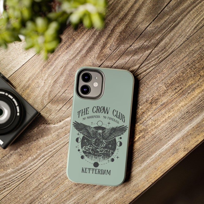 May include: A green phone case with a black and white illustration of a crow with the text "The Crow Club No Mourners No Funerals Ketterdam".