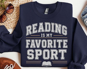 May include: Navy blue sweatshirt featuring the text "READING IS MY FAVORITE SPORT" in white, collegiate-style lettering. An open book graphic is below the text. A brown shoe and sunglasses are visible near the folded sweatshirt.