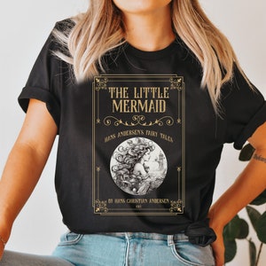 Pu&ograve; includere: Maglietta nera con una grafica dorata di una copertina di libro con un'illustrazione di sirena e il testo "THE LITTLE MERMAID", "HANS ANDERSEN'S FAIRY TALES", "BY HANS CHRISTIAN ANDERSEN", e "1913".