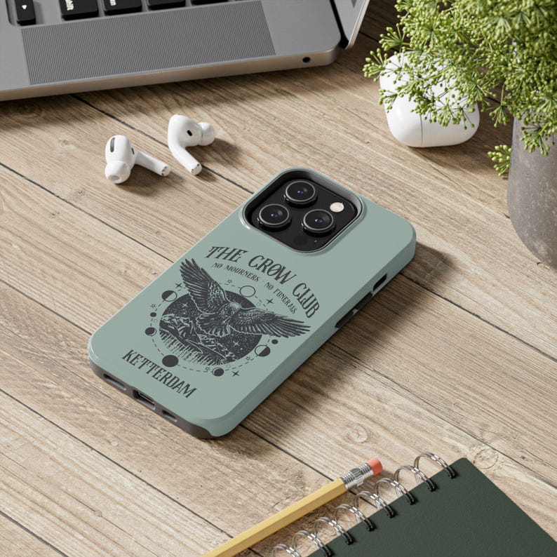 May include: A green phone case with a black and white illustration of a crow with the text "The Crow Club - No Mourners - No Funerals - Ketterdam" printed on it.