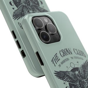 May include: A sage green phone case with a black and white illustration of a crow with the text "The Crow Club No Mourners No Funerals".