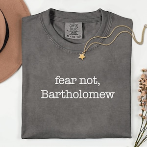 May include: A dark gray t-shirt with the words "fear not, Bartholomew" in white. A gold star pendant necklace and a brown hat are also in the image. The shirt has a Comfort Colors tag.