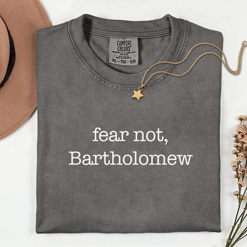 May include: A dark gray t-shirt with the words "fear not, Bartholomew" in white. A gold star pendant necklace and a brown hat are also in the image. The shirt has a Comfort Colors tag.