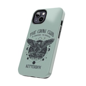 May include: A light blue phone case with a black and white illustration of a crow with the text "The Crow Club No Mourners No Funerals Ketterdam".