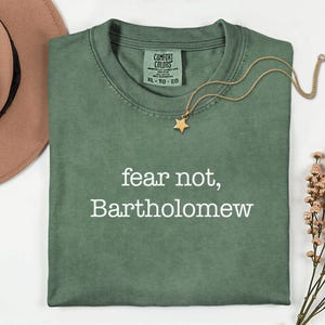 May include: Sage green Comfort Colors t-shirt with the text "fear not, Bartholomew" in white. A gold star pendant necklace and a brown hat are also visible. The shirt is folded neatly on a white surface.