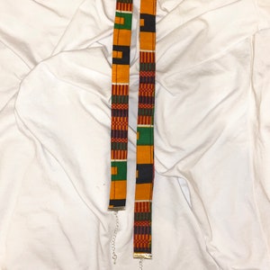 Puede incluir: Dos gargantillas de tela con un estampado africano colorido en tonos de amarillo, verde, rojo y negro. Las gargantillas est&aacute;n unidas a cadenas plateadas.