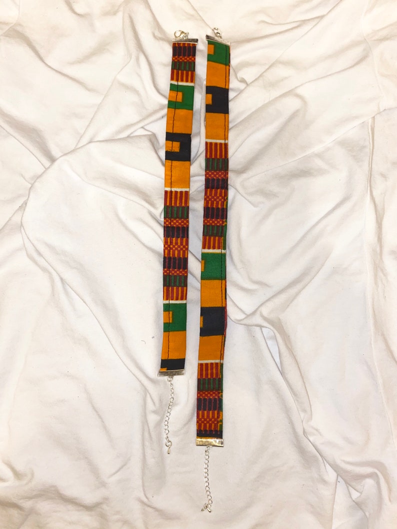 Puede incluir: Dos gargantillas de tela con un estampado africano colorido en tonos de amarillo, verde, rojo y negro. Las gargantillas est&aacute;n unidas a cadenas plateadas.