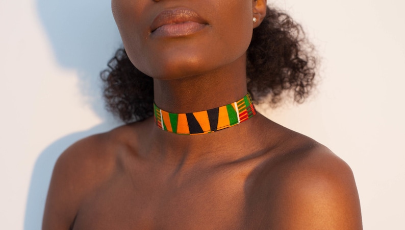 Puede incluir: Primer plano de una persona que lleva un collar de tela colorido con rayas negras, naranjas, verdes y rojas. El collar est&aacute; hecho de una tela tejida con un patr&oacute;n geom&eacute;trico.