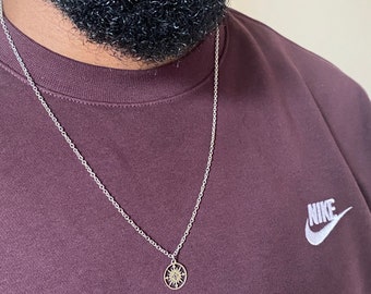 Könnte beinhalten: Eine silberne Kettenkette mit einem kleinen, runden Anhänger mit einem Sonnendesign. Der Anhänger ist goldfarben und hat ein detailliertes Design. Die Kette wird von einer Person getragen, die ein maroonfarbenes Sweatshirt mit einem weißen Nike-Logo trägt.