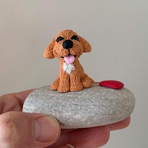 May include: A small, brown clay figurine of a dog sitting on a grey rock with a red heart-shaped object beside it.