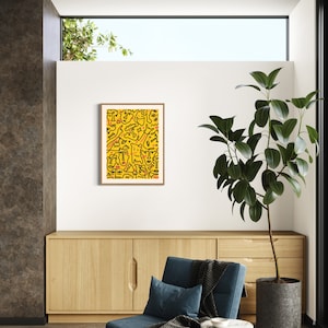 May include: A framed abstract art print with a yellow background and black line drawings of faces. The print is hanging on a white wall in a living room setting with a blue armchair and a potted plant.