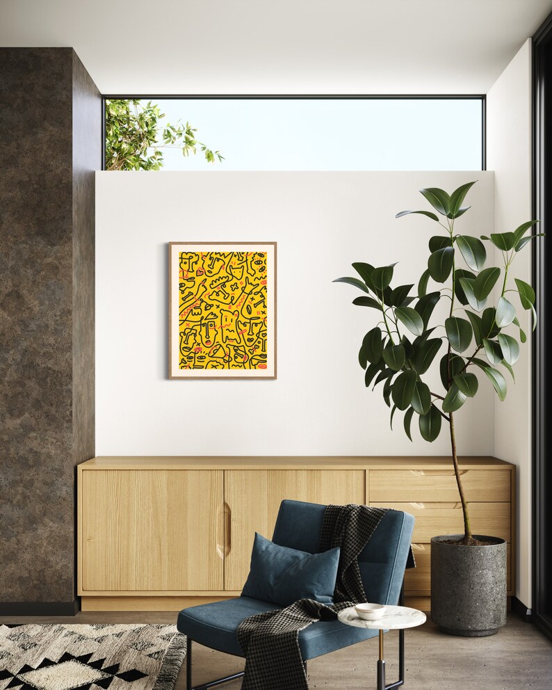 May include: A framed abstract art print with a yellow background and black line drawings of faces. The print is hanging on a white wall in a living room setting with a blue armchair and a potted plant.