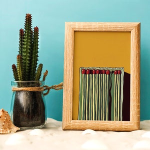 May include: A framed print of a minimalist illustration of a box of matches with red match heads. The background is a yellow color. The print is in a wooden frame.