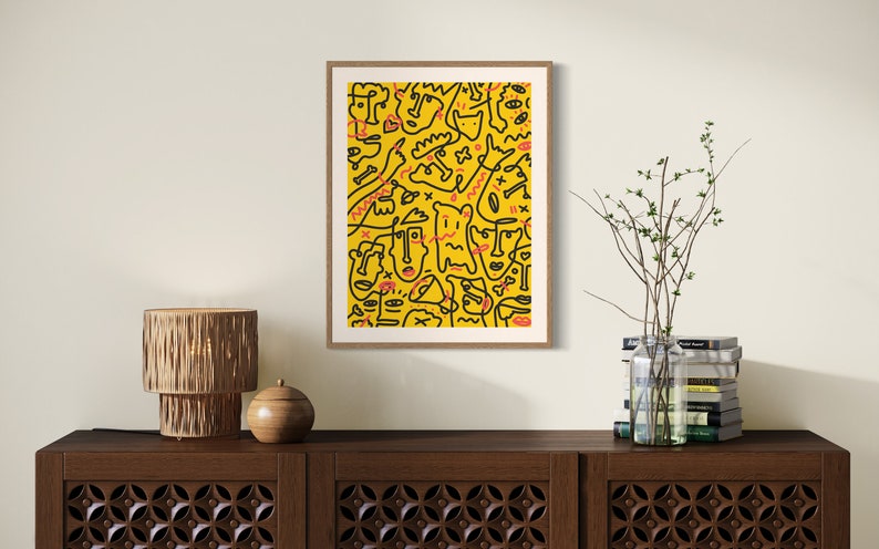 May include: A framed print with a yellow background and black and red abstract line drawings. The print is hanging on a white wall above a wooden cabinet.