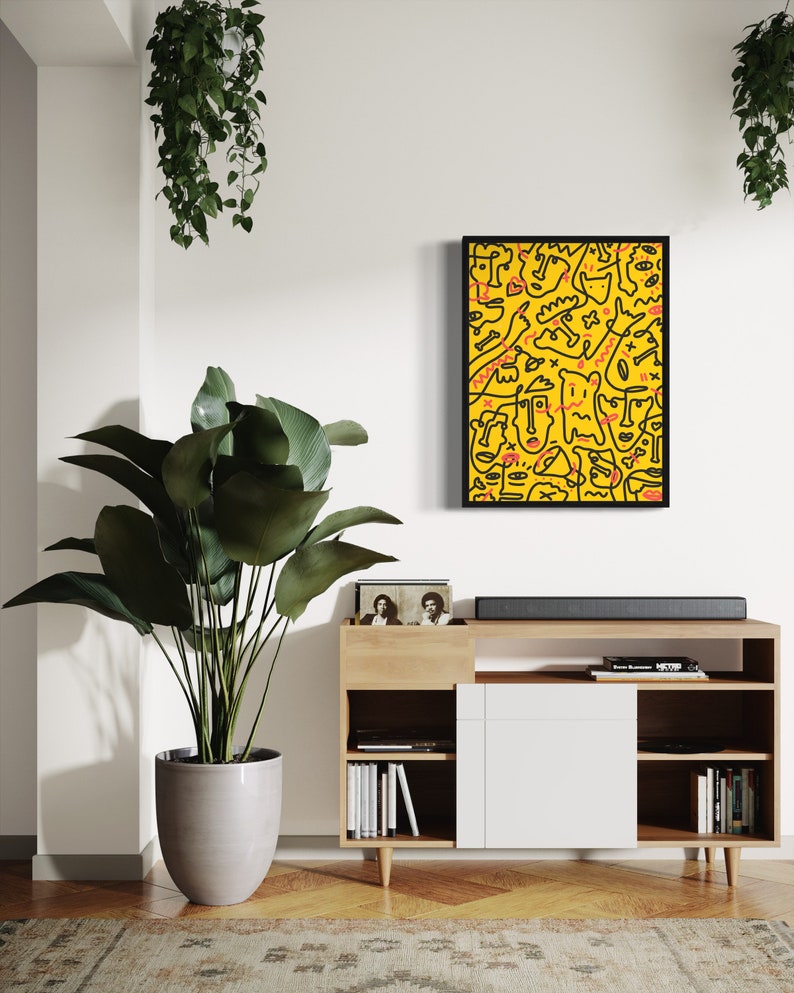 May include: A black framed print of a yellow background with black line drawings of faces and abstract shapes. The print is hanging on a white wall above a wooden shelf with books and a record player.