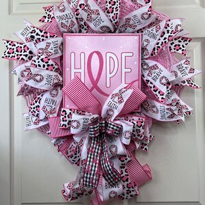May include: Pink and white wreath with a pink ribbon and the word "HOPE" on a sign. The wreath is decorated with leopard print ribbon and pink and white ribbon with the words "faith", "hope", "love", "brave", and "strong".