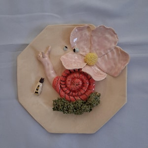 May include: A ceramic wall hanging featuring a whimsical snail with a red shell and a pink flower. The snail has a raised arm and is surrounded by green foliage. A small, striped insect is also present.