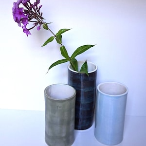 May include: Three cylindrical ceramic vases with white interiors. One vase is sage green, one is dark navy blue, and one is light blue. A single stem of purple phlox flowers is placed inside the dark navy blue vase, extending upwards.