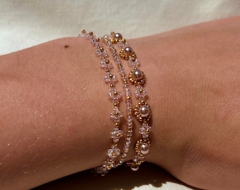 May include: A set of four delicate bracelets featuring small, white beads and gold accents. The bracelets are layered on a person's wrist, creating a stylish and feminine look.