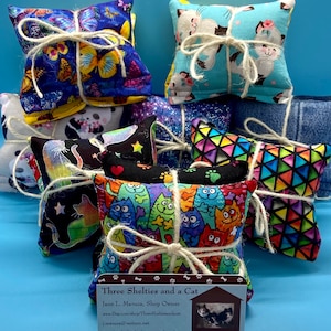May include: A stack of small, square pet toys made of colorful fabric, tied with twine. The fabric features various patterns, including butterflies, cats, and geometric designs. A business card with the text "Three Shelties and a Cat" is visible.