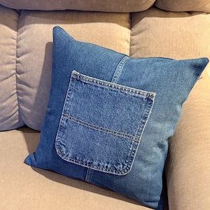 May include: A denim pillow with a pocket detail. The pillow is made from blue jean material and has a rectangular pocket sewn onto the front. The pillow is resting on a beige sofa.