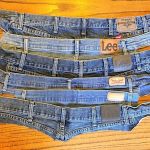 May include: An assortment of denim jean waistbands in various shades of blue. The waistbands are from different brands, including Wrangler, Lee, and Levi's, each with its brand label. The jeans are laid out on a wooden surface.